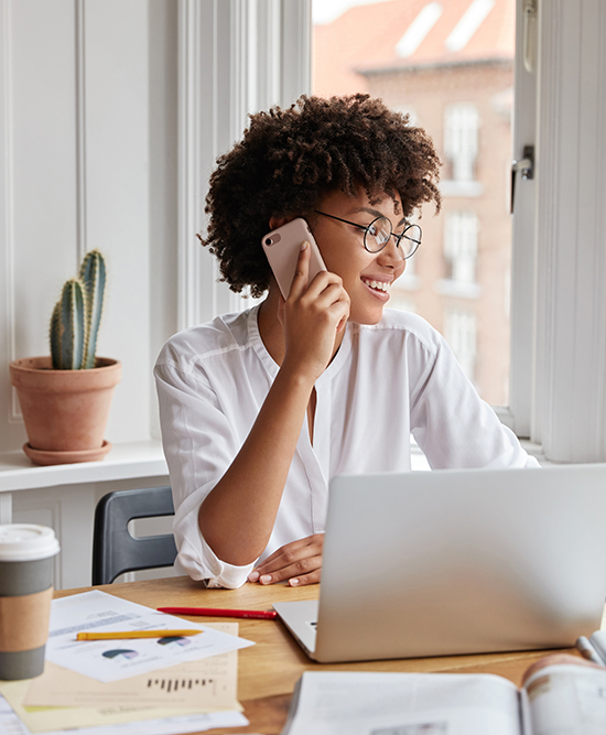 Vertical shot of cheerful woman bookkeeper talks on mobile phone, has positive expression, dressed in white stylish blouse, works with laptop computer, has work break, drinks hot aromatic beverage